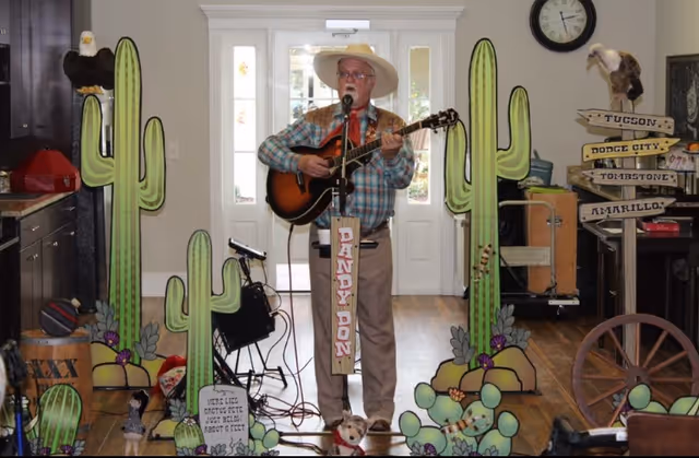 An elderly man wearing a cowboy hat and plaid shirt plays an acoustic guitar and sings into a microphone in a room decorated with large cardboard cutouts of cacti and desert plants. There are props including a wooden signpost with city names, a wagon wheel, and a small dog figurine. The man stands in front of a white door with glass panels, and a clock is visible on the wall behind him.