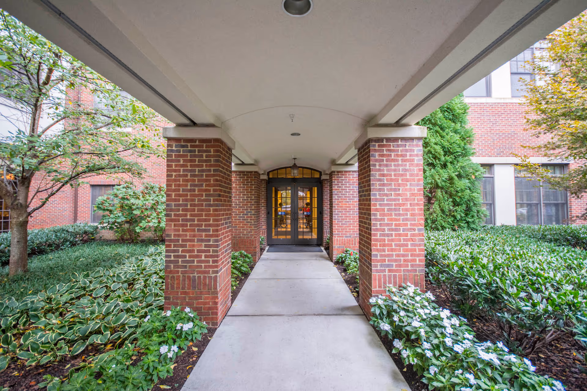 Covered walkway with brick pillars leading to glass double doors of a building, surrounded by green shrubs, trees, and white flowers.