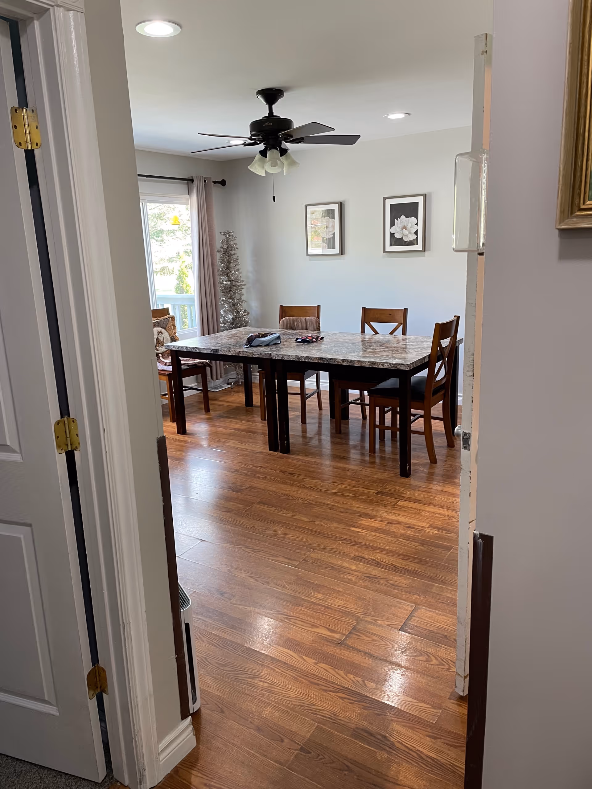 View through an open doorway into a dining room with a rectangular table and six wooden chairs. The room has wooden flooring, a ceiling fan with lights, two framed floral pictures on the wall, and a window with curtains letting in natural light. A decorative silver Christmas tree is in the corner.