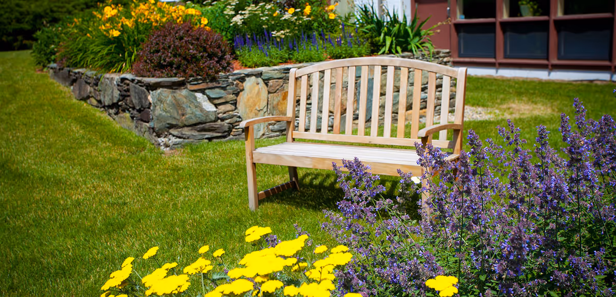 A wooden bench placed on a well-maintained green lawn surrounded by colorful flowers including yellow and purple blooms, with a stone flower bed and part of a building visible in the background.
