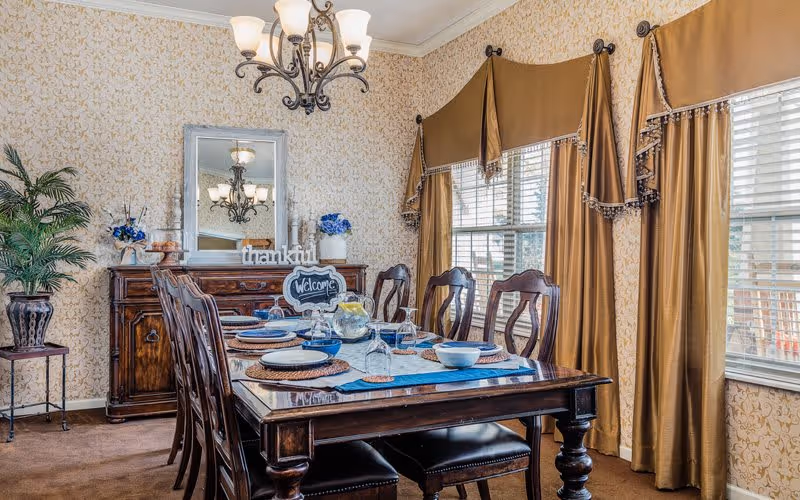 A traditional dining room with a dark wooden dining table set for six, featuring blue and white dishware and placemats. The room has patterned wallpaper, a chandelier overhead, and large windows with gold curtains. A wooden sideboard with decorative items, including a 'thankful' sign and a 'welcome' chalkboard, is against the wall.