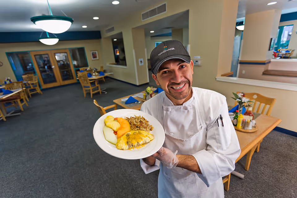 A smiling chef wearing a white uniform and black cap holds a plate of food with an omelette, rice, and sliced fruit in a dining room with wooden tables and chairs set with blue napkins.