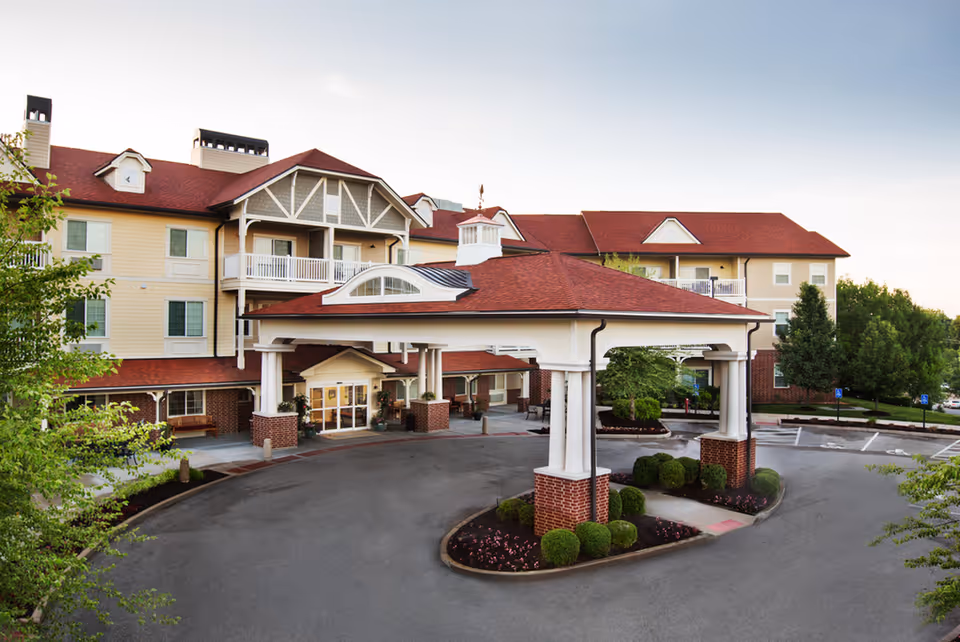 Exterior view of Crestview Senior Living facility showing a large covered entrance with white columns and brick bases, surrounded by landscaped bushes and trees, with a multi-story building featuring beige siding and red roofs in the background.