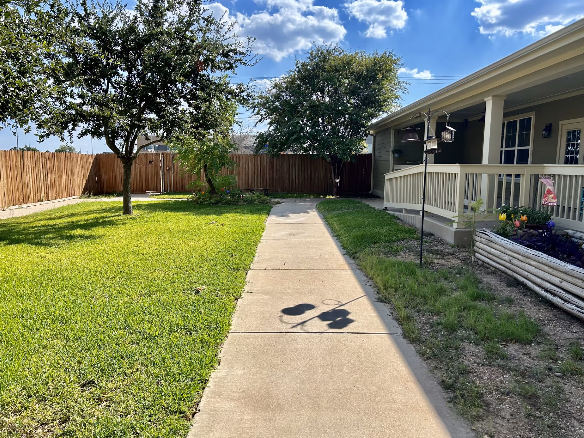 Outdoor area of Mercy House Temple featuring a concrete walkway flanked by green grass, trees, and a wooden fence. On the right side, there is a building with a covered porch, railings, and a garden bed with flowers and a bird feeder.