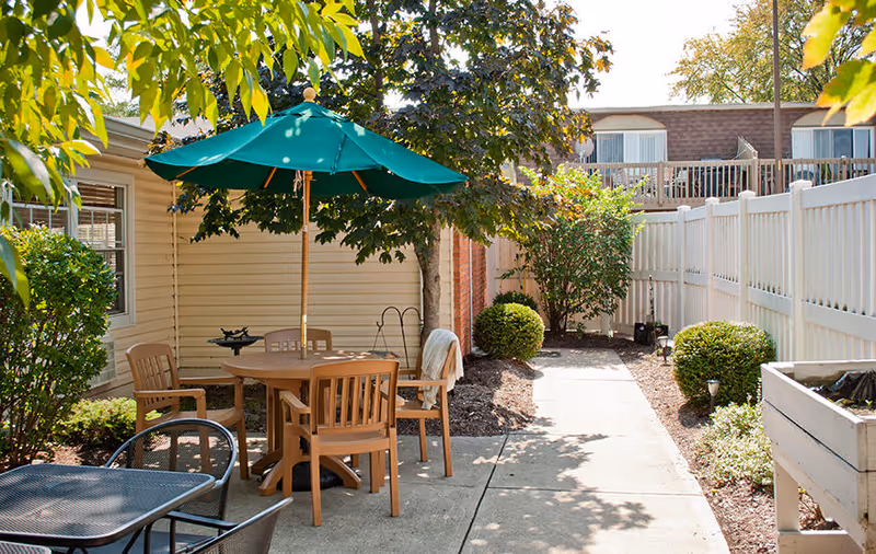 Outdoor patio area with a round wooden table and four chairs under a green umbrella, surrounded by trees and bushes. A concrete pathway runs alongside the seating area, with a white fence on one side and a building on the other.