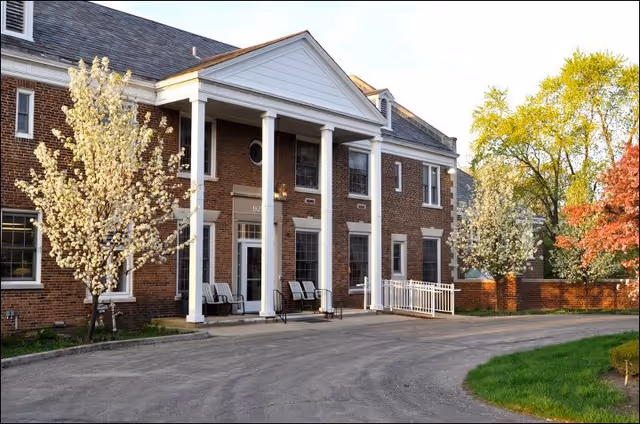 Front entrance of a brick building with white columns, chairs on the porch, blossoming trees and a curved driveway.
