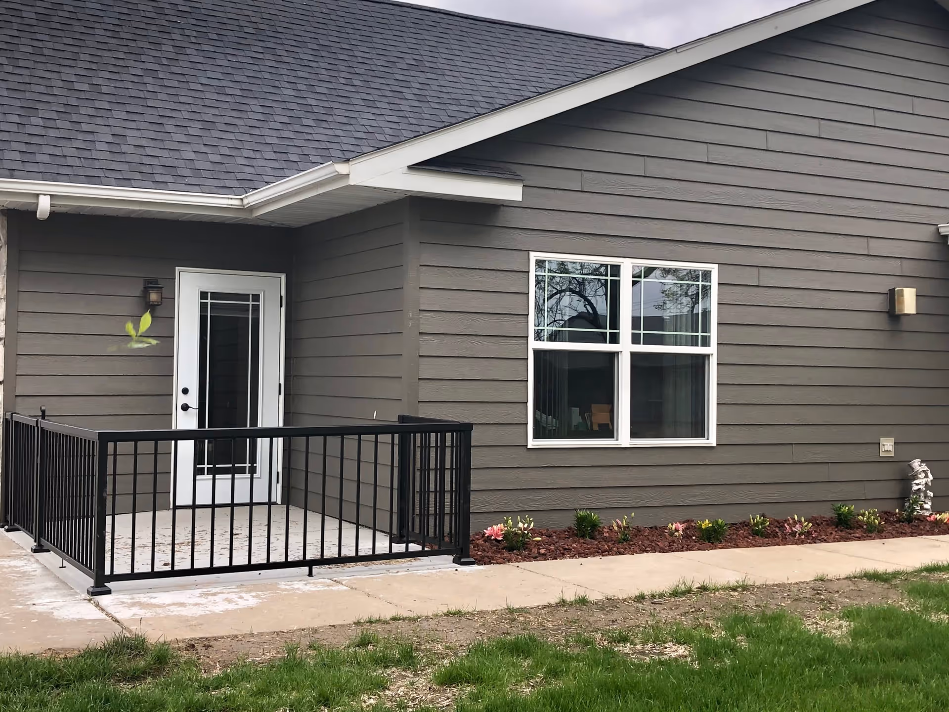 Exterior view of a gray building with a small fenced patio area, a white door with glass panels, a window with white trim, and a flower bed with small plants along the side. The building has a shingled roof and a concrete walkway in front.