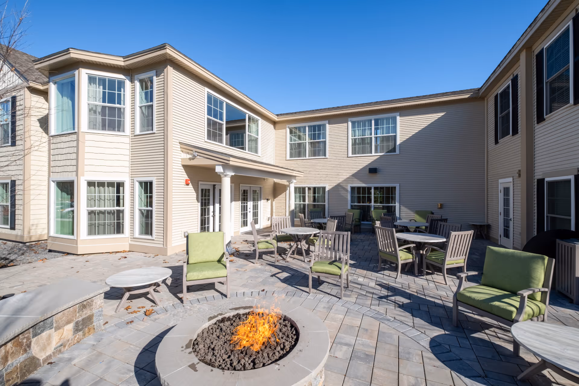 Outdoor patio area at Benchmark Senior Living at Nashua Crossings with multiple tables and chairs featuring green cushions arranged around a central fire pit. The patio is surrounded by a beige two-story building with many windows under a clear blue sky.