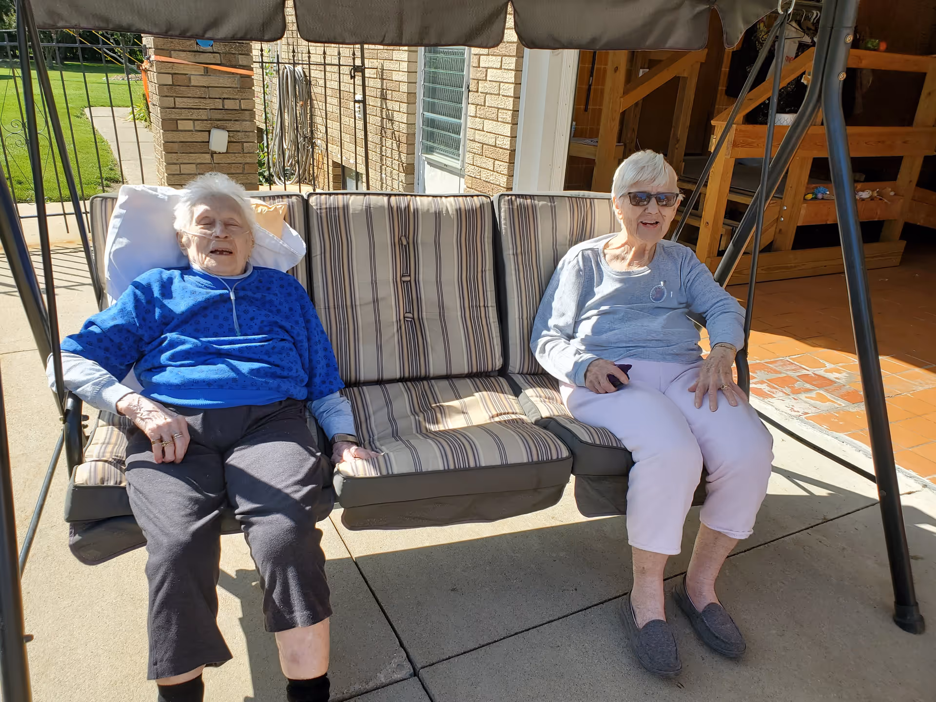 Two elderly women sitting on a cushioned outdoor swing bench under a canopy. One woman is wearing a blue jacket and black pants, and the other is wearing a gray long-sleeve shirt and light pink pants with sunglasses. They are outside near a brick building with a concrete patio and some greenery visible in the background.