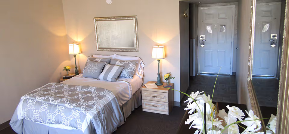 A neatly made bed with patterned bedding and multiple pillows in a cozy bedroom. Two bedside tables each have a lamp and a small flower arrangement. A large mirror is mounted on the wall above the bed. There is a dresser with white flowers in the foreground and a door visible in the background.