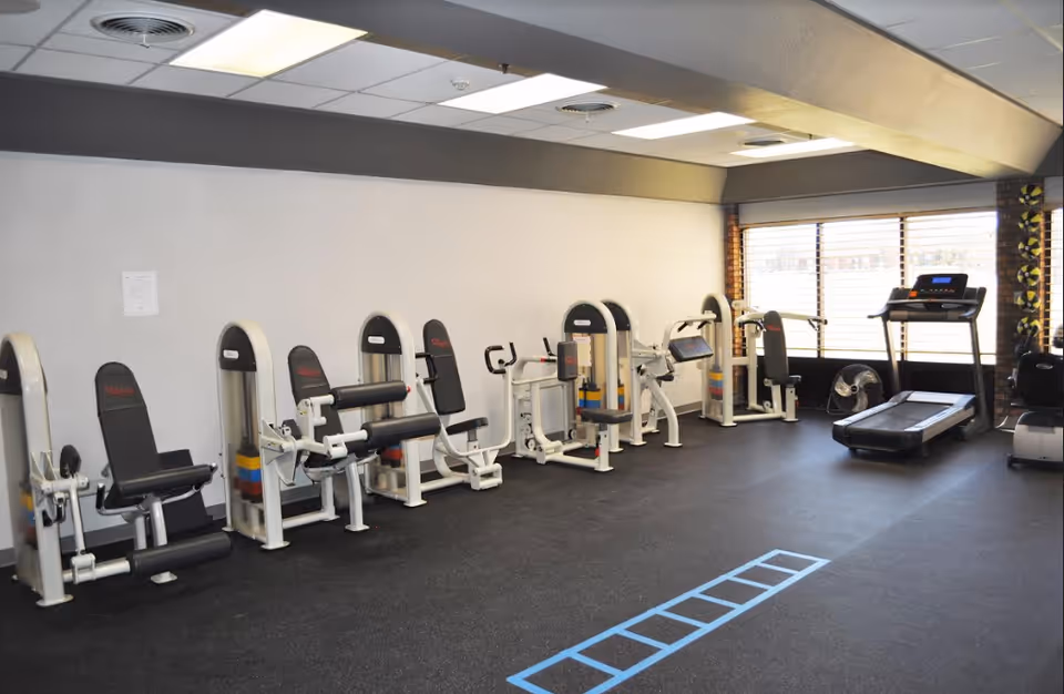 Interior view of a fitness room with various exercise machines lined up against a white wall, including leg press and arm curl machines, and a treadmill near a large window with blinds. The floor has a black rubber mat with a blue agility ladder pattern.