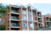 Exterior view of a multi-story brick residential building with balconies and white columns, surrounded by some greenery under a clear sky.