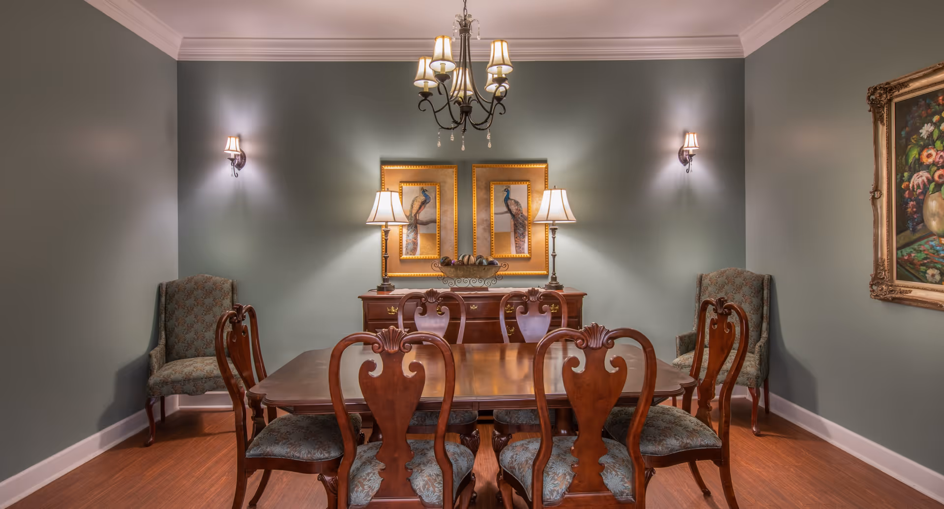 A traditional dining room with a wooden dining table surrounded by six ornate chairs with patterned upholstery. Two armchairs are placed against the walls on either side. The walls are painted a muted green color and decorated with framed artwork featuring peacocks and a floral painting. The room is softly lit by a chandelier hanging from the ceiling, two wall sconces, and two table lamps on a wooden sideboard.