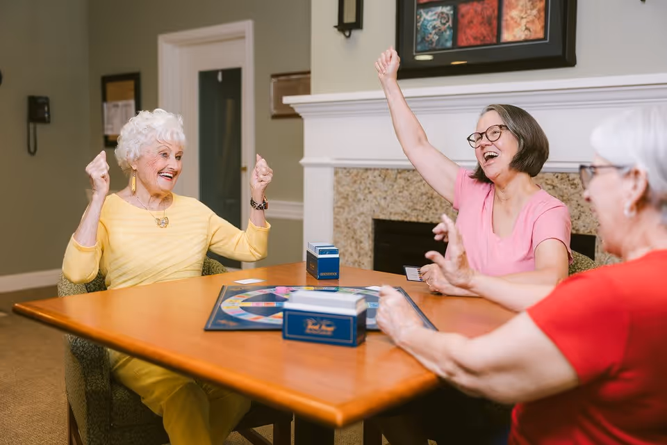 Three elderly women sitting around a wooden table playing a board game in a cozy room with a fireplace and framed artwork on the wall. Two women are raising their arms in celebration, smiling and enjoying the game.
