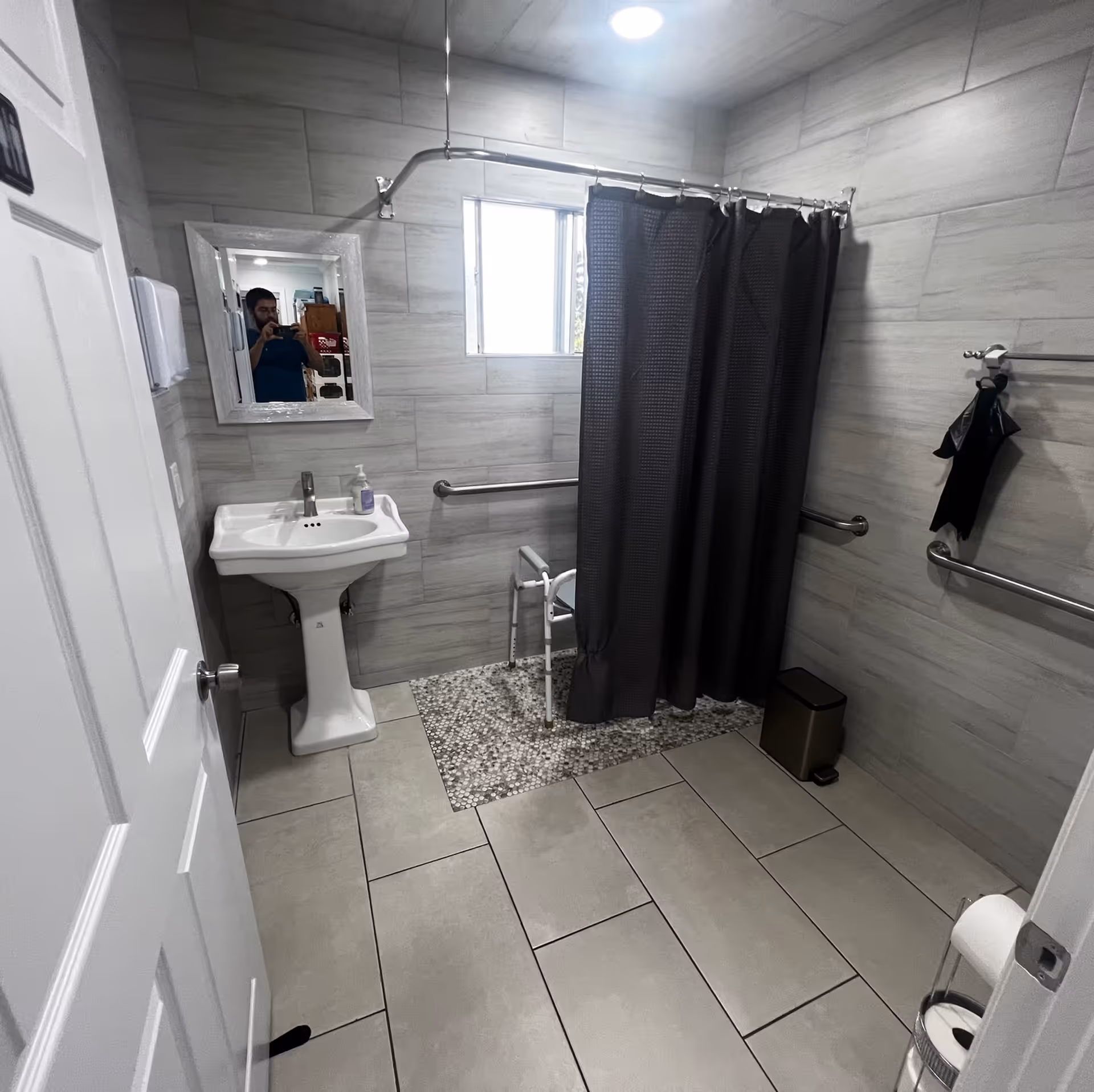 Accessible bathroom with a pedestal sink, grab bars, a walk-in shower with a dark curtain and shower chair, and tiled floors.