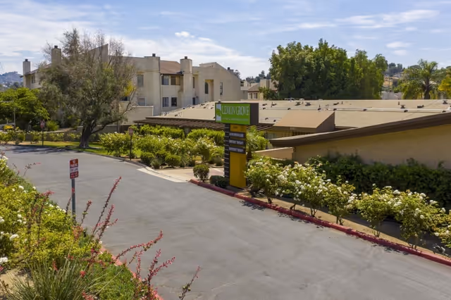 Exterior view of Lemon Grove Care And Rehabilitation Center showing a driveway lined with bushes and flowers, a green and yellow facility sign, and multiple beige buildings with trees in the background under a clear sky.