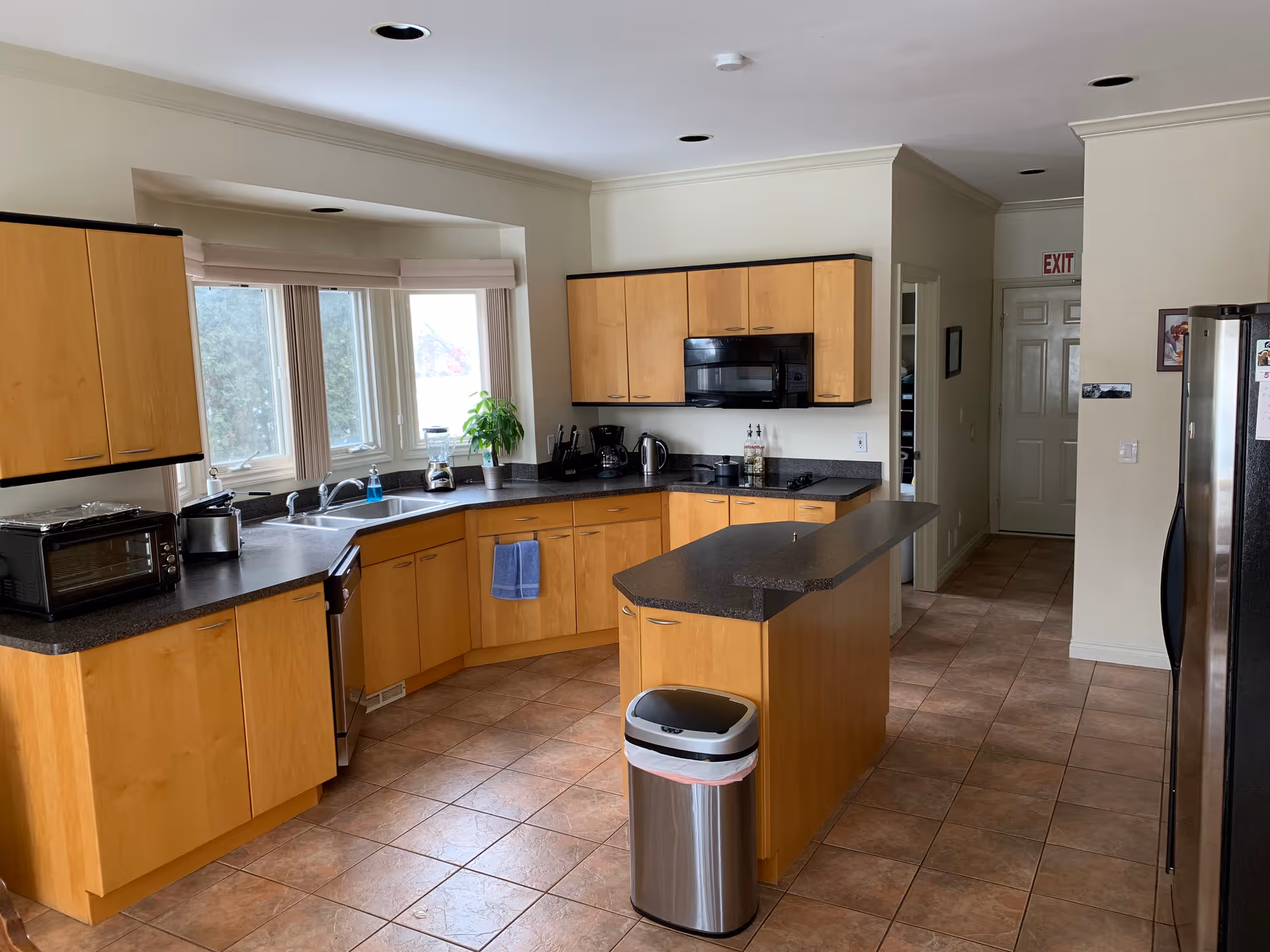Spacious kitchen with light wood cabinets, a center island, stainless appliances and trash can, and tiled floor.