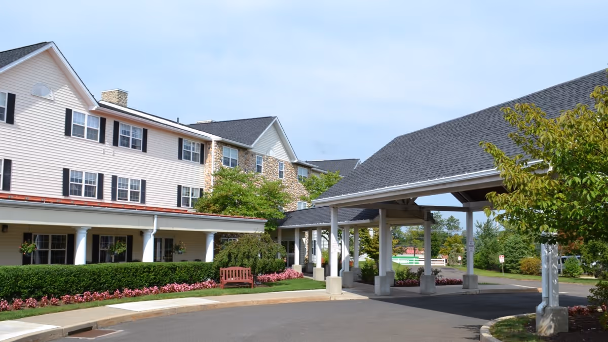 Front exterior of a multi-story senior living building with a covered porte-cochere, landscaped flower beds, and a bench.