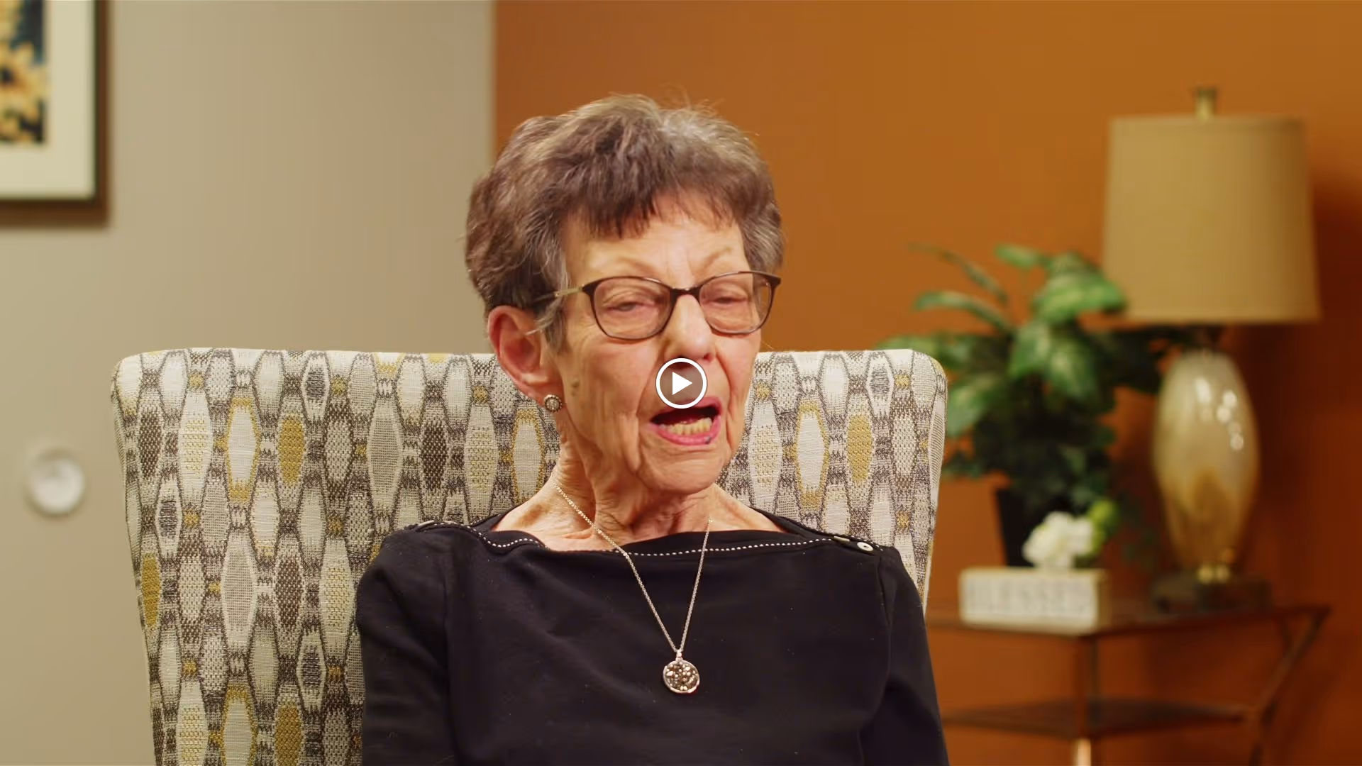 An elderly woman with short gray hair and glasses is sitting in a patterned armchair. She is wearing a black top and a necklace with a circular pendant. Behind her, there is a table with a lamp, a plant, and a decorative sign that says 'BLESSED'. The wall behind her is painted in two tones, beige and orange.