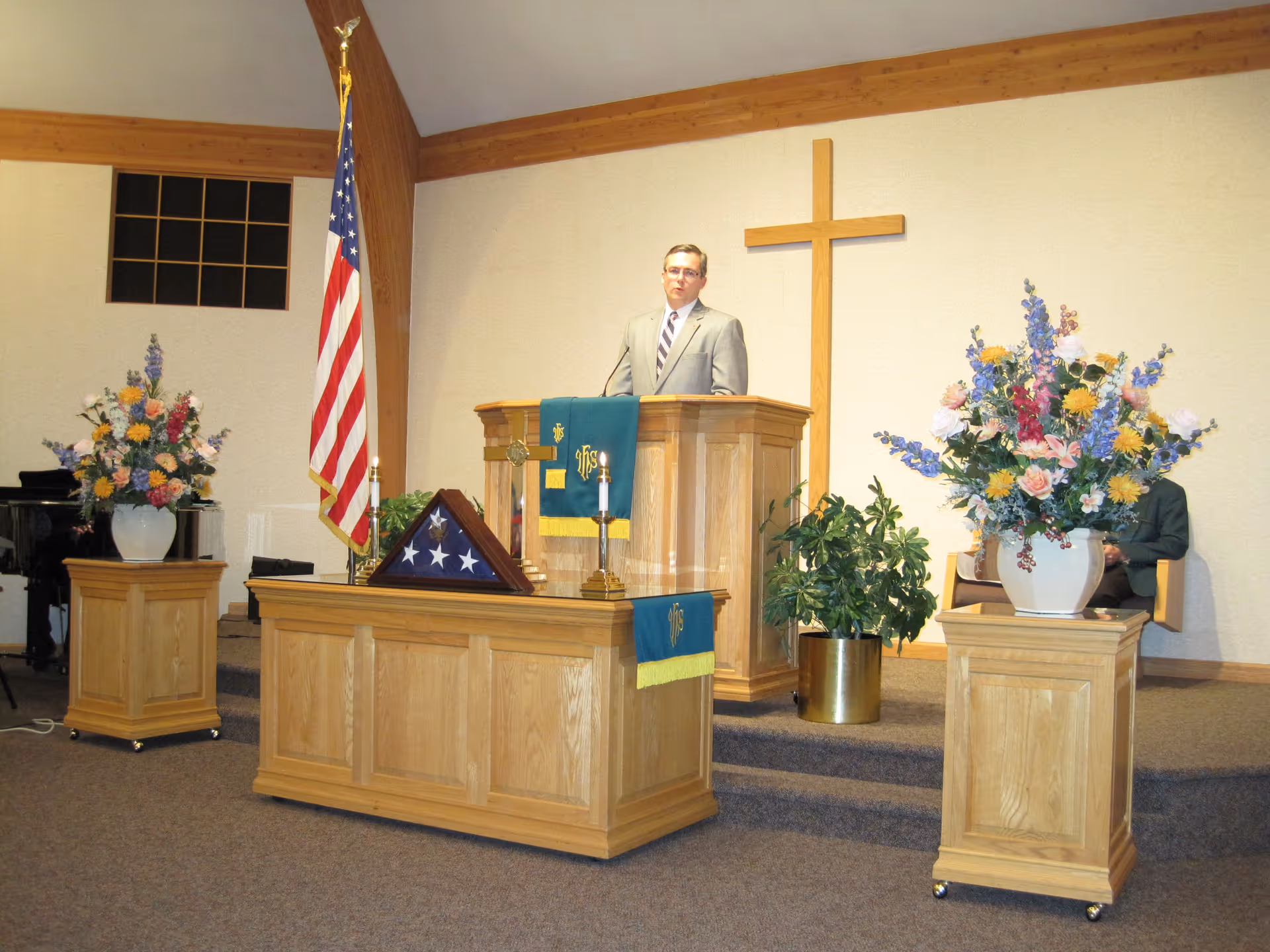 A man in a suit stands behind a wooden pulpit in a chapel. Behind him is a large wooden cross mounted on the wall. The pulpit and a smaller wooden table in front of it are decorated with green cloths featuring religious symbols. An American flag stands to the left of the pulpit. Two large floral arrangements in white vases are placed on wooden pedestals on either side of the pulpit. There is a folded American flag displayed on the smaller table, along with two candles. A green potted plant is visible behind the pulpit.