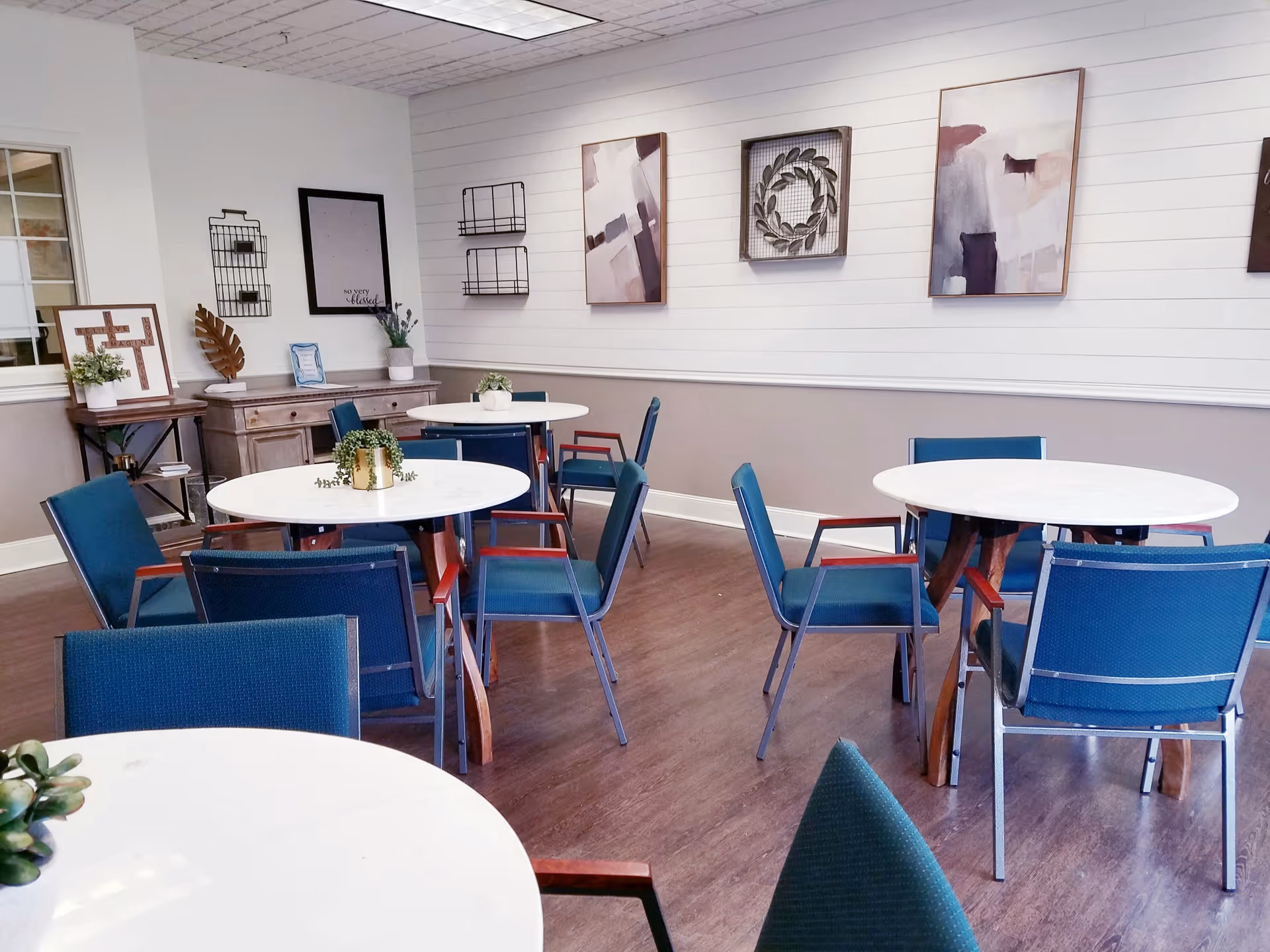 A bright and clean dining room with round white tables and blue chairs. The walls are decorated with abstract paintings and a decorative wreath. There are small potted plants on the tables and a sideboard with additional decor against the wall.