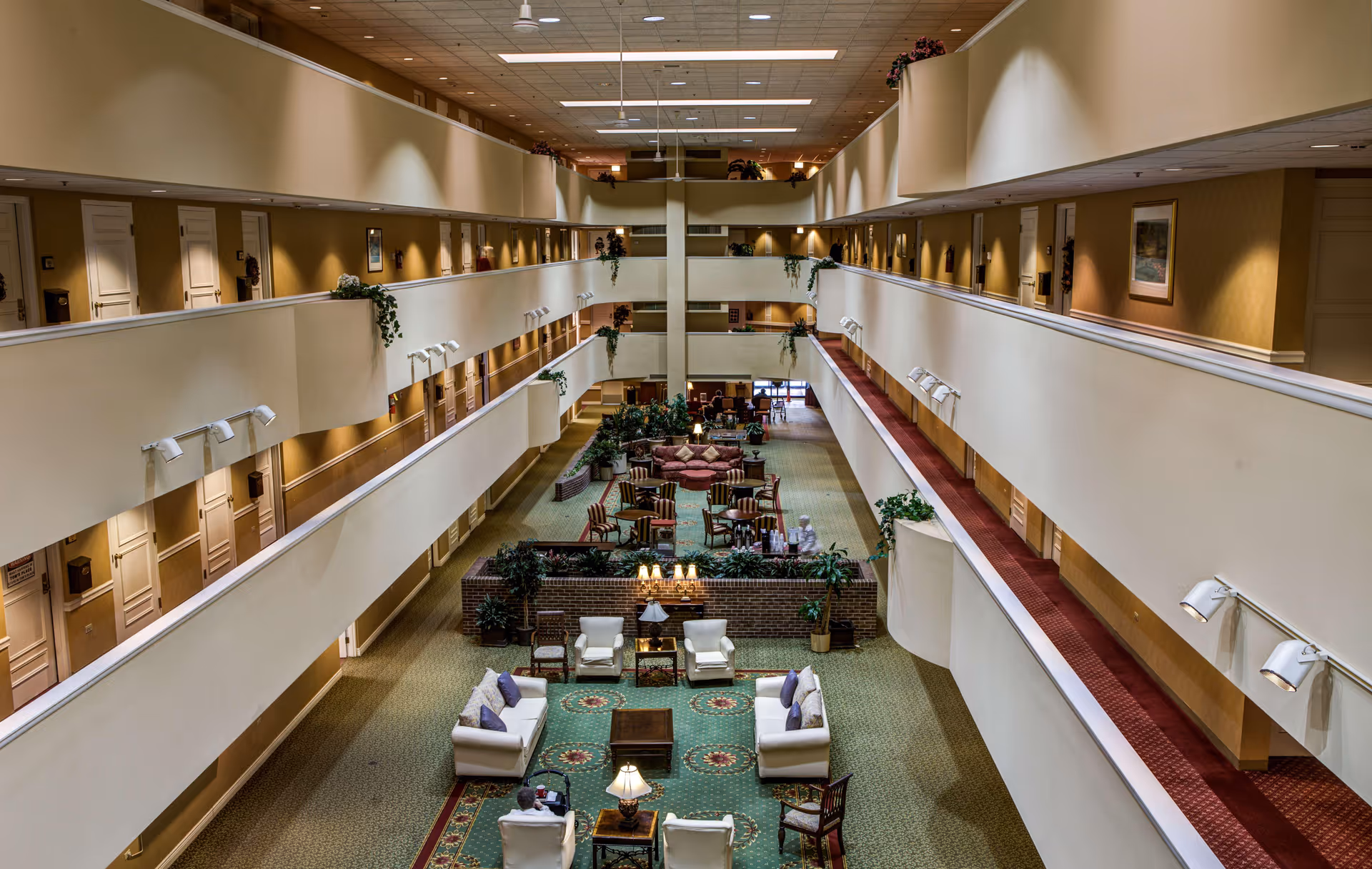 Interior view of a multi-level senior living facility with a central open atrium. The atrium features seating areas with sofas, armchairs, and tables arranged on patterned carpets. The upper levels have hallways with doors leading to rooms, decorated with framed pictures and plants along the railings.