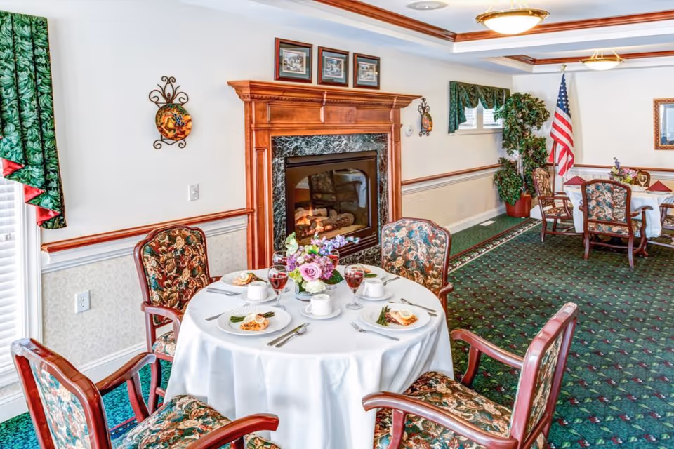 Dining room with a round table set for a meal, floral centerpiece, patterned chairs, a fireplace, and green carpet.