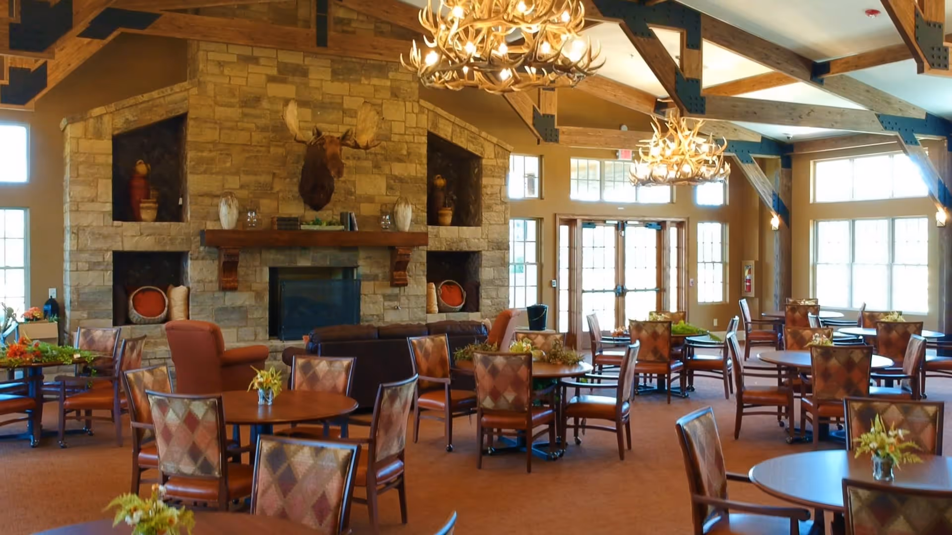 A spacious dining area with multiple round tables and cushioned chairs arranged neatly. The room features a large stone fireplace with a mounted moose head above it, decorative shelves with vases, and large windows allowing natural light to fill the space. The ceiling has exposed wooden beams and antler chandeliers hanging down, creating a warm and rustic atmosphere.