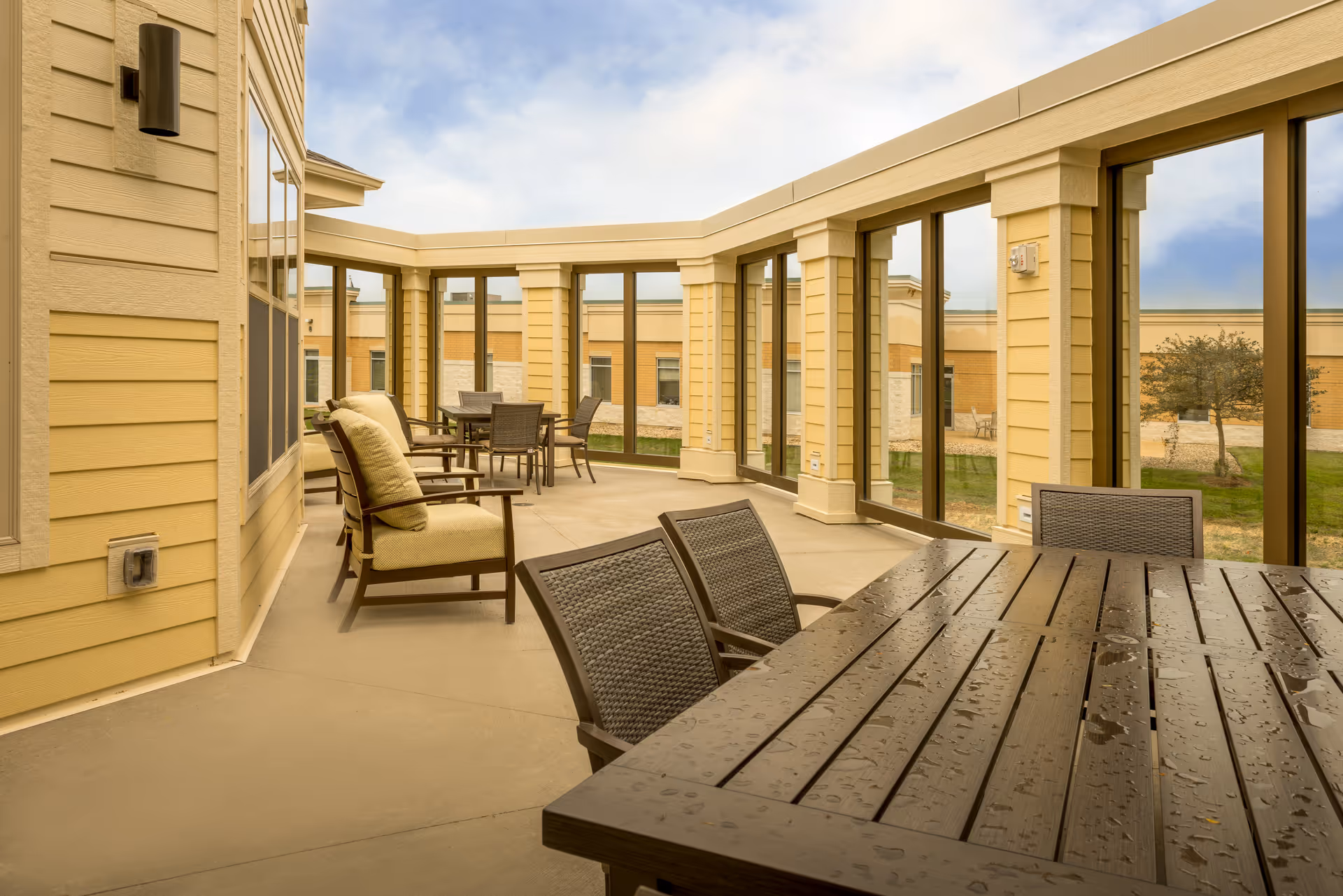 A covered outdoor patio area at Brightstar Senior Living Of Waunakee with cushioned chairs and tables. The patio is enclosed with large glass windows, allowing views of the outside garden and building exterior. The table in the foreground has water droplets on its surface, indicating recent rain.