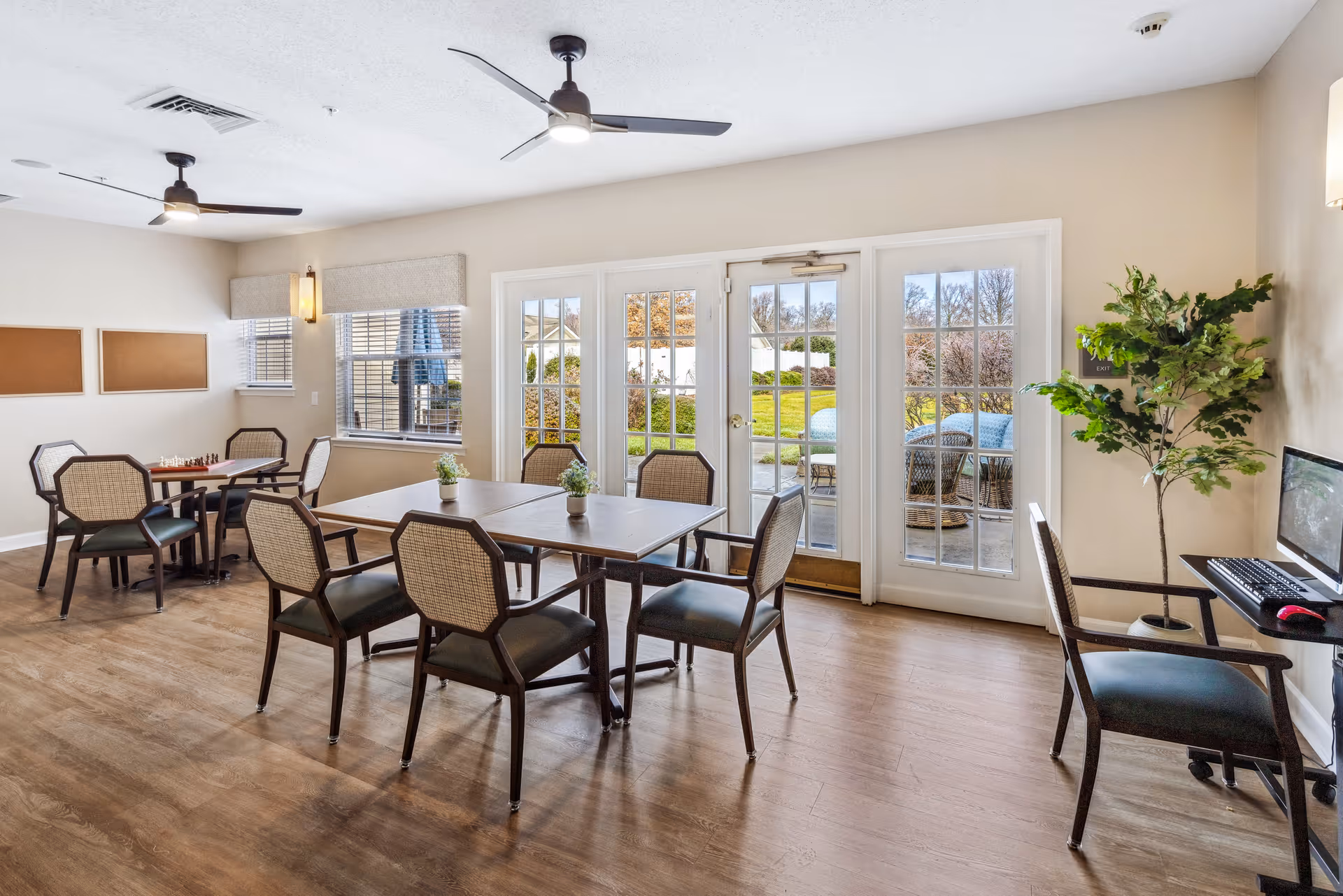 A bright and spacious common area with wooden flooring, featuring several tables and chairs arranged for group seating. Large windows and glass doors provide a view of a green outdoor patio area with additional seating. The room includes ceiling fans, a potted plant, and a computer workstation with a chair on the right side.