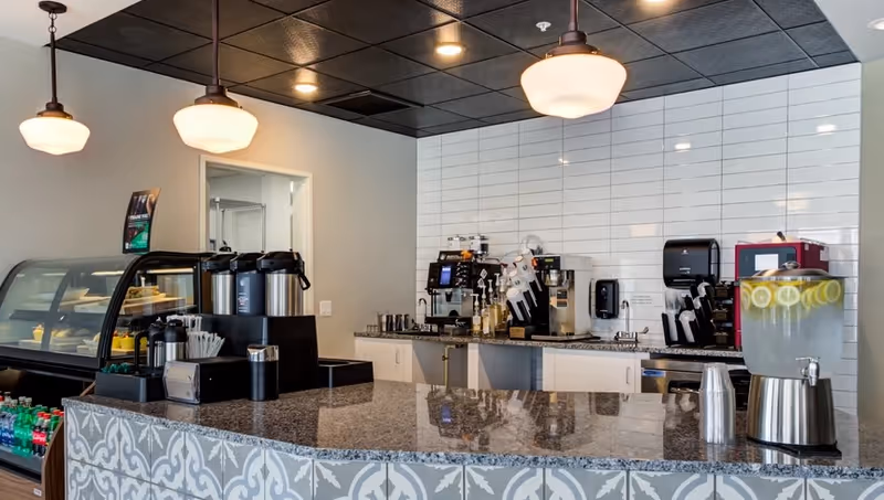 A modern coffee and beverage station with a granite countertop, several coffee dispensers, a water dispenser with lemon slices, and various coffee machines against a white tiled wall. There are hanging pendant lights above the counter and a refrigerated display case with bottled drinks to the side.