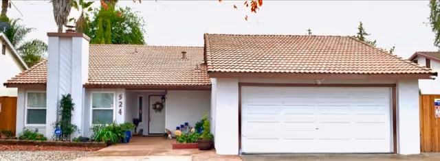 Front exterior view of a single-story house with a tiled roof, white walls, a chimney, and a two-car garage. The house has a small porch area with plants and a wreath on the front door. The address number 524 is visible near the entrance.
