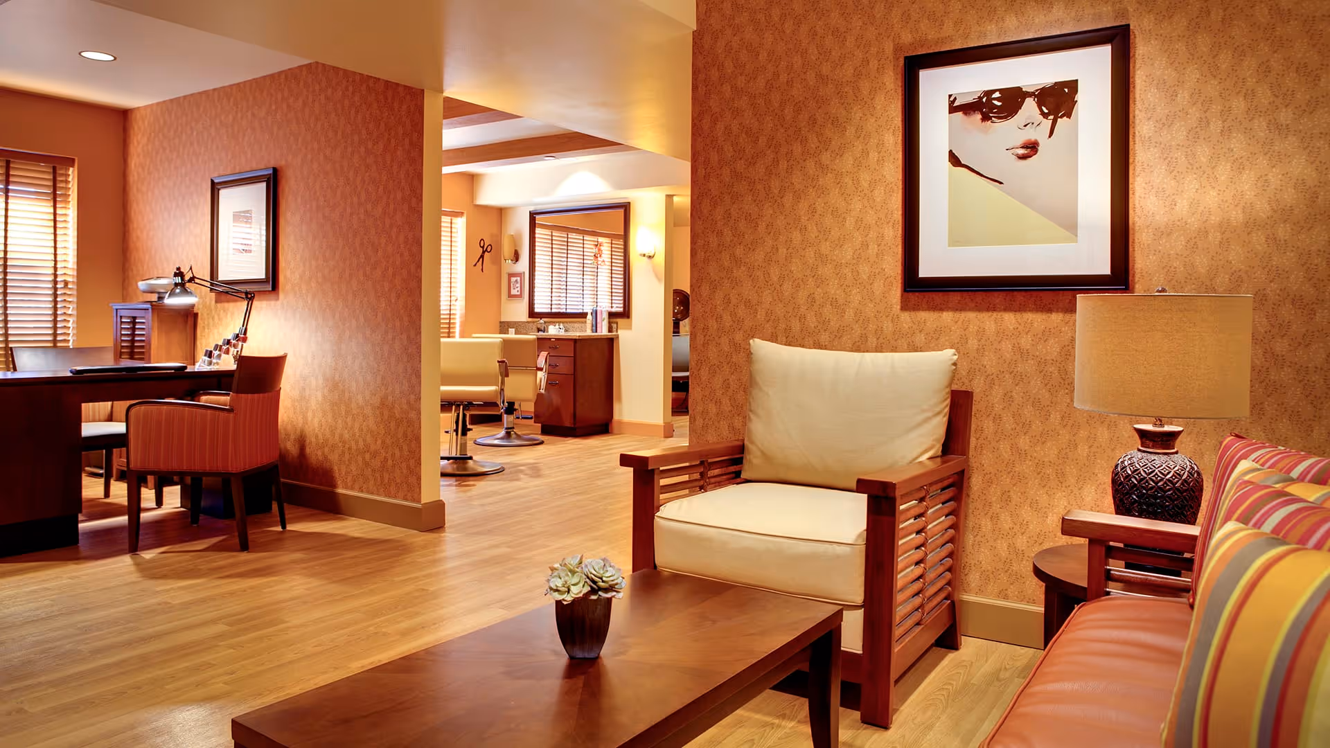 A cozy interior space with warm lighting featuring a wooden armchair with cream cushions, a wooden coffee table with a small potted plant, a striped sofa with colorful cushions, and a side table with a decorative lamp. In the background, there is a desk with chairs and a lamp, and further back, a salon area with styling chairs and mirrors. The walls have a patterned wallpaper and framed artwork.