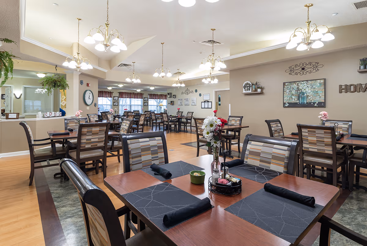 A spacious dining room in a senior living facility with multiple wooden tables and chairs arranged neatly. Each table has black placemats, rolled black napkins, and a centerpiece with flowers. The room is well-lit with several hanging chandeliers and has beige walls decorated with artwork and shelves. Large windows with valances allow natural light to enter the room.