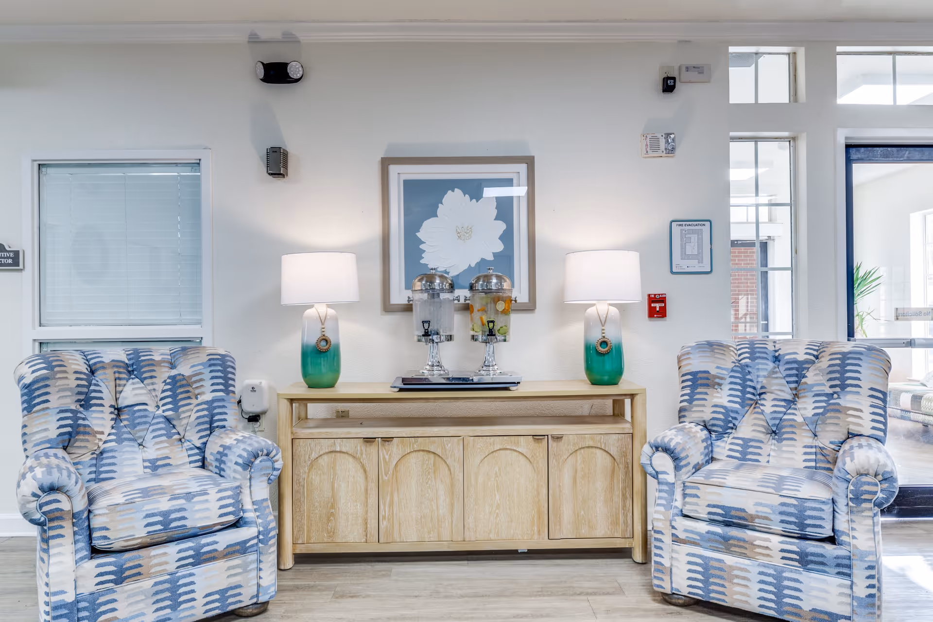 A cozy seating area in a senior living facility featuring two patterned armchairs with blue, beige, and white colors. Between the chairs is a wooden sideboard with two green and white table lamps and two beverage dispensers. Above the sideboard is a framed picture of a white flower on a blue background. The room has light-colored walls and flooring, with a window and glass door visible on the sides.