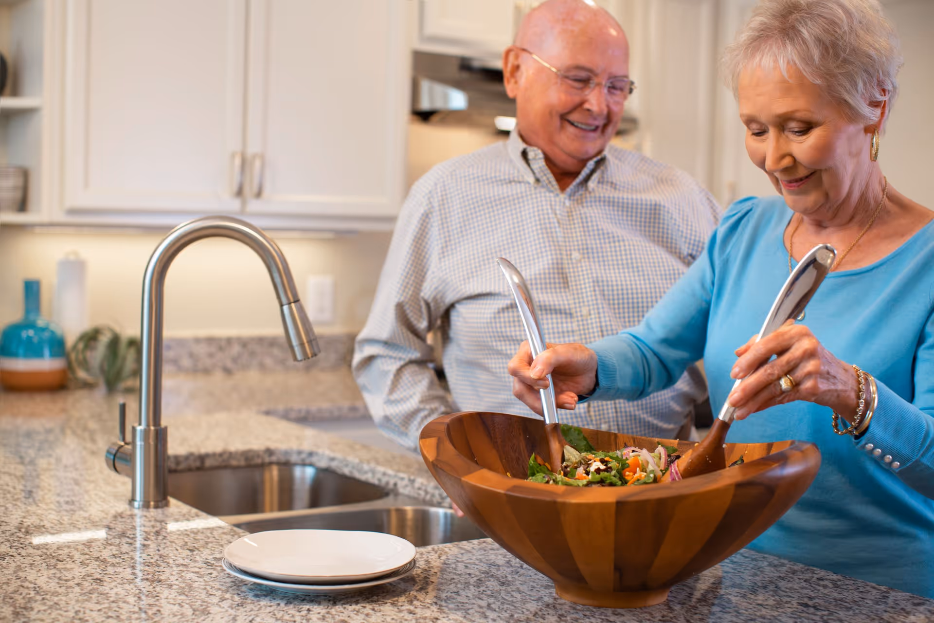 An elderly woman and man are in a kitchen. The woman is mixing a salad in a large wooden bowl using salad servers, while the man stands behind her smiling. The kitchen has a granite countertop with a sink and faucet, white cabinets, and some decorative items in the background.