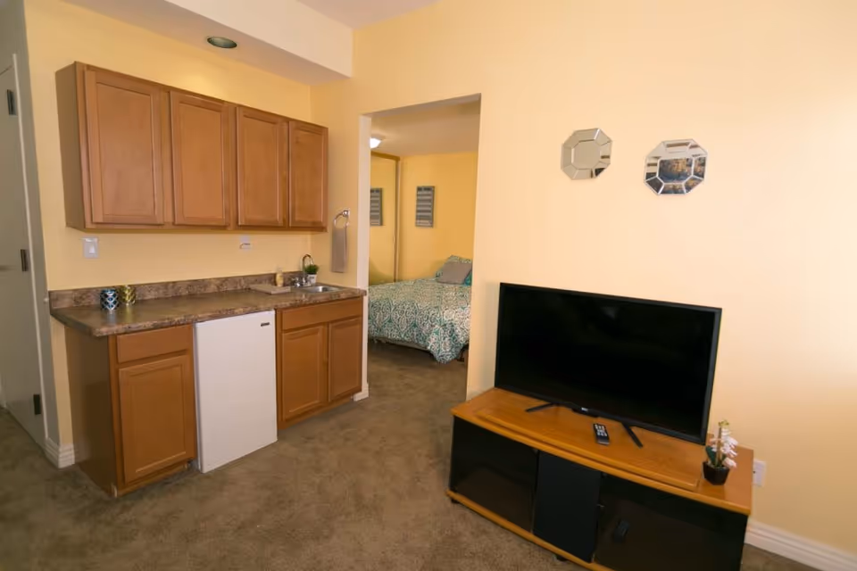 Interior view of a senior living facility room with a small kitchenette featuring wooden cabinets, a countertop, a sink, and a mini refrigerator. Adjacent to the kitchenette is a living area with a flat-screen TV on a wooden TV stand. In the background, a bedroom with a bed covered in a blue patterned bedspread is visible through an open doorway. The walls are painted light yellow and the floor is carpeted.