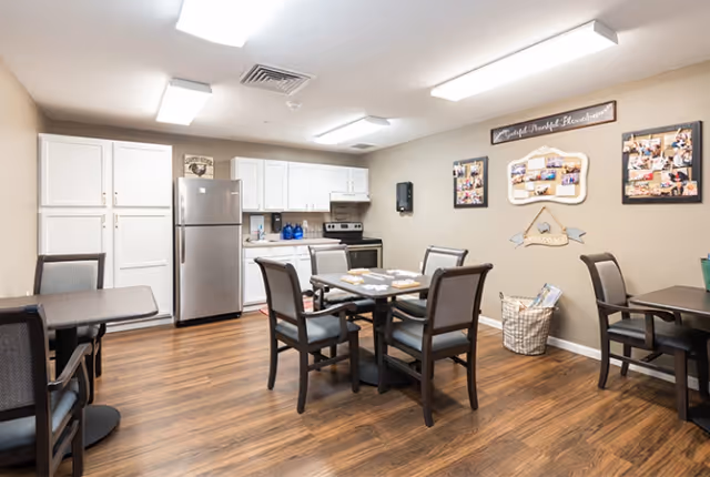 A bright and clean communal dining area with a small kitchen in the background. The kitchen features white cabinets, a stainless steel refrigerator, and a stove. The dining area has several tables with chairs arranged around them. The walls are decorated with framed photos and a wooden sign. The floor is wood laminate.
