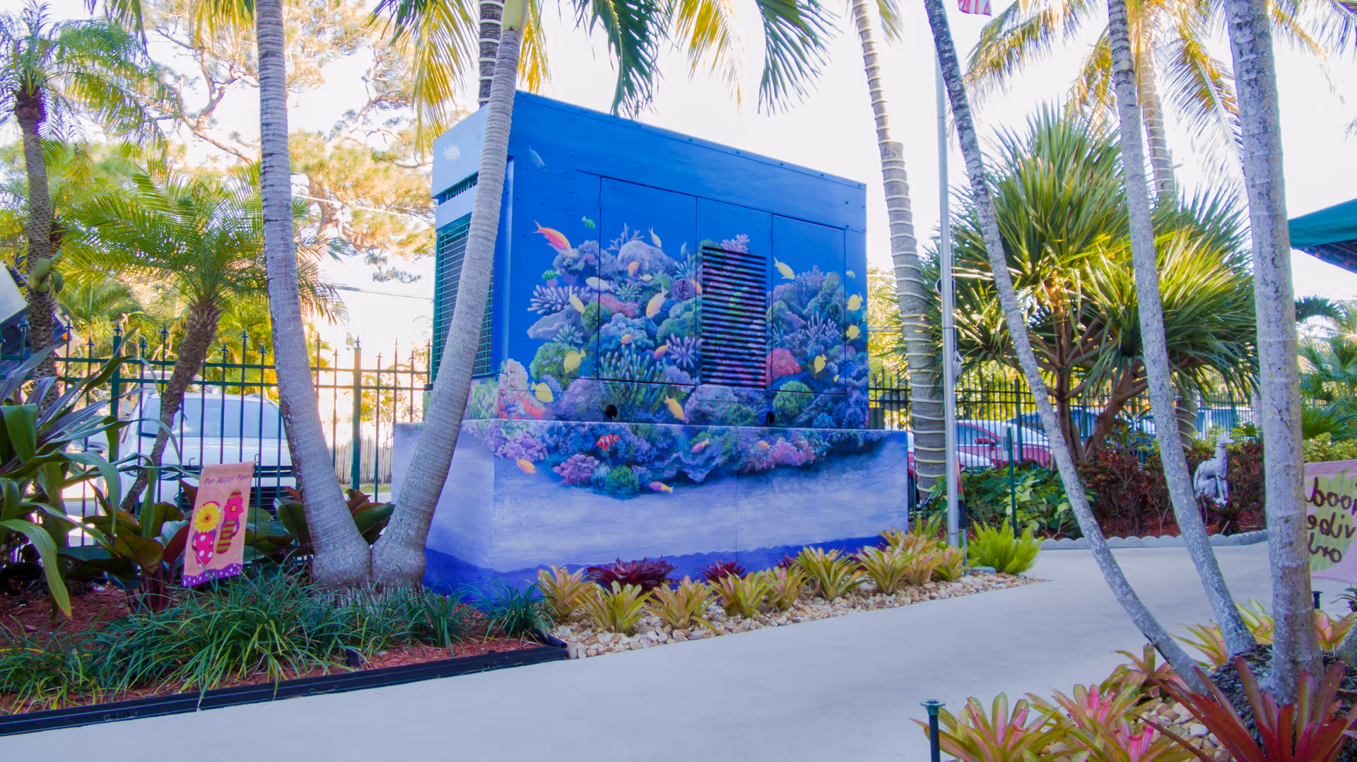 Outdoor area with tropical plants and palm trees surrounding a large blue utility box painted with a colorful underwater coral reef mural. There is a paved walkway and a black metal fence in the background.