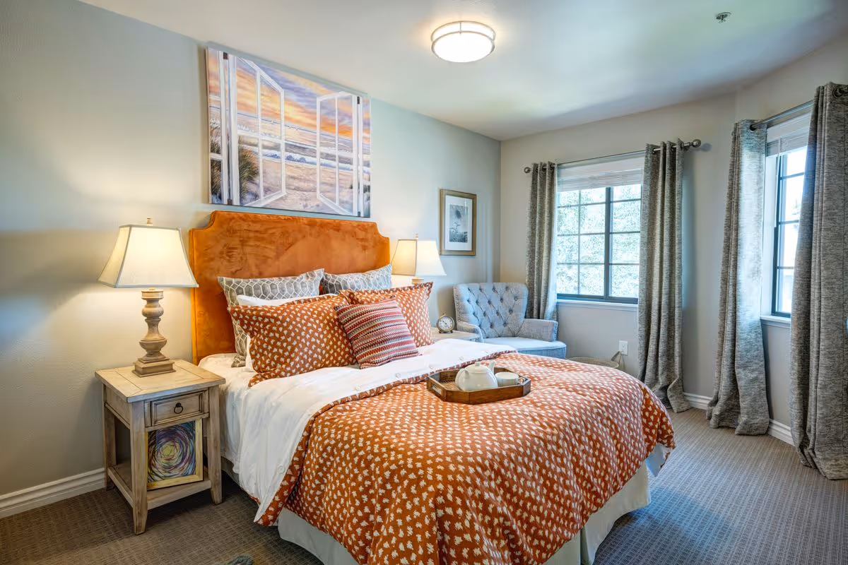 A cozy bedroom with a large bed featuring an orange patterned comforter and multiple pillows. Two bedside tables with lamps flank the bed. A cushioned armchair sits near two windows with gray curtains, allowing natural light to fill the room. A framed picture and a painting of an open window overlooking a beach scene hang on the walls.
