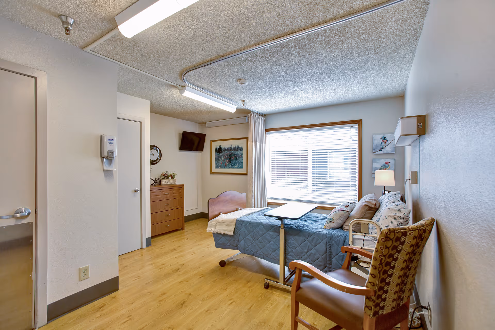 A bright senior living bedroom with a single hospital-style bed, overbed table, armchair, dresser, and a large window with blinds.