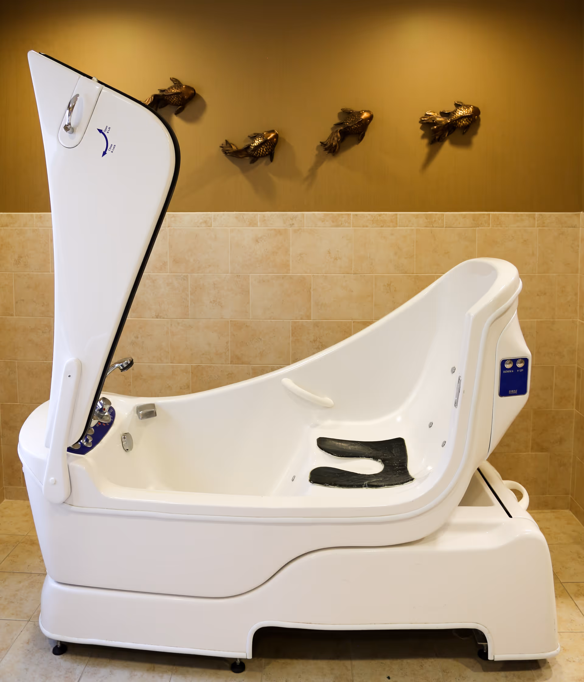 A white assisted bathing tub with its lid open sits against a tiled wall decorated with three metal fish.