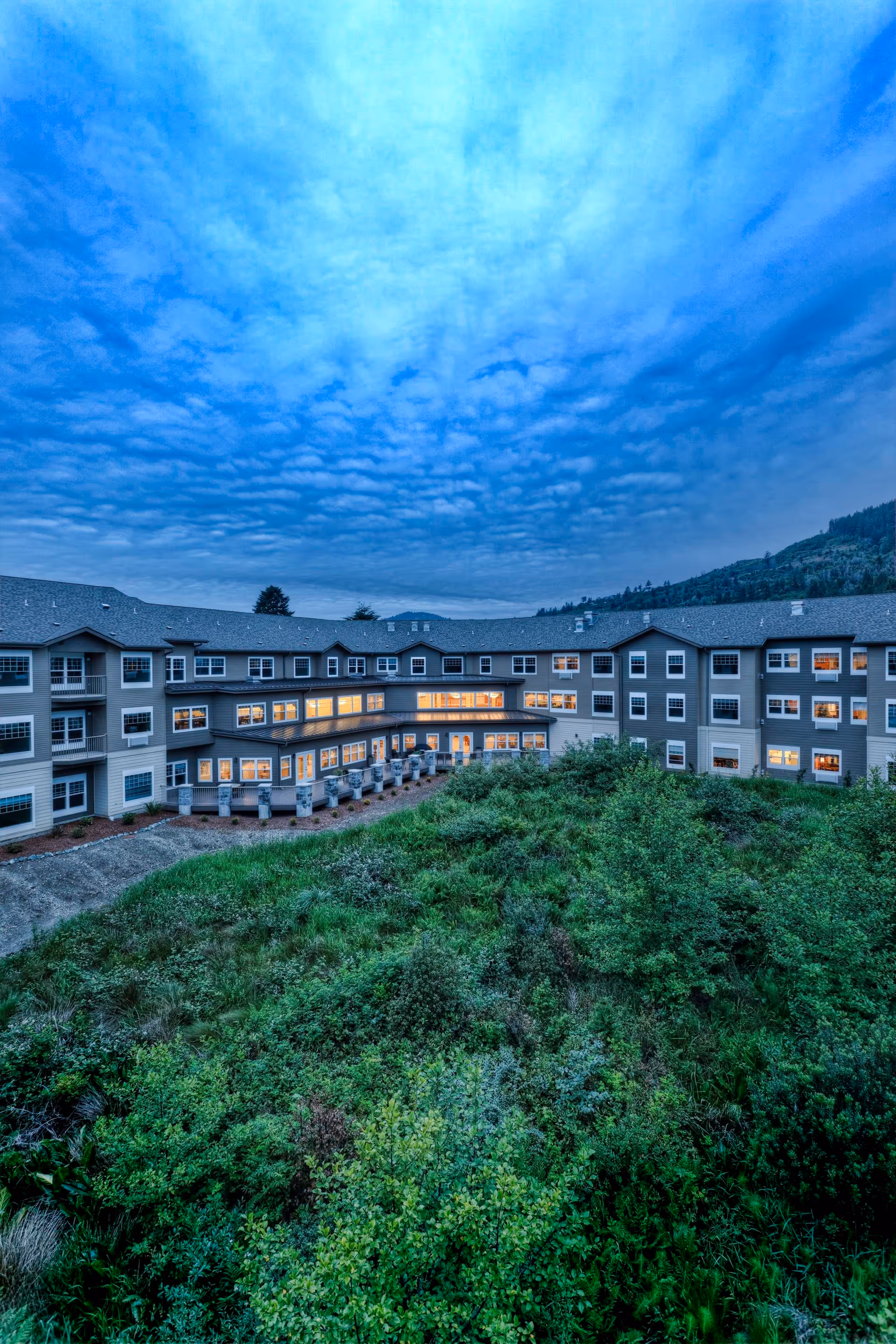 Exterior view of a large senior living community building at dusk with many windows lit up, surrounded by green shrubbery and hills under a cloudy blue sky.