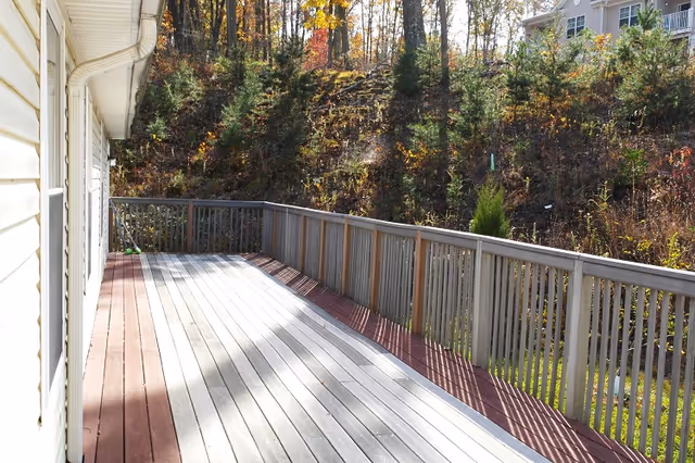 A wooden outdoor deck attached to a light-colored house, with a railing along the edge. The deck overlooks a wooded area with trees and shrubs, and sunlight filters through the trees casting shadows on the deck.
