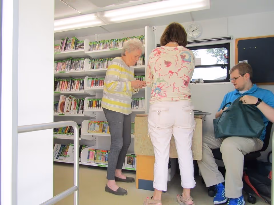 Three people inside a small library-like room with shelves of books, two standing at a counter and one seated holding a bag.