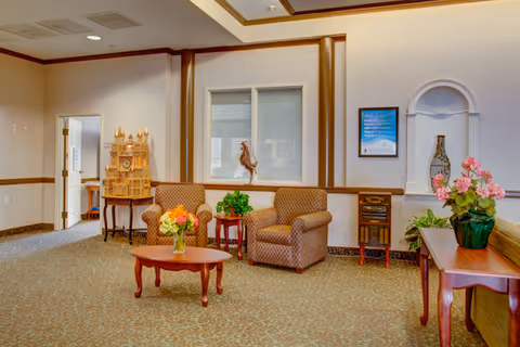 A cozy sitting area in a senior living facility with two patterned armchairs, a wooden coffee table with a vase of flowers, a small side table with a plant, and decorative items including a wooden model and a rooster figurine. The walls are light-colored with wooden trim, and there is a framed sign and a decorative niche with a vase on the wall.