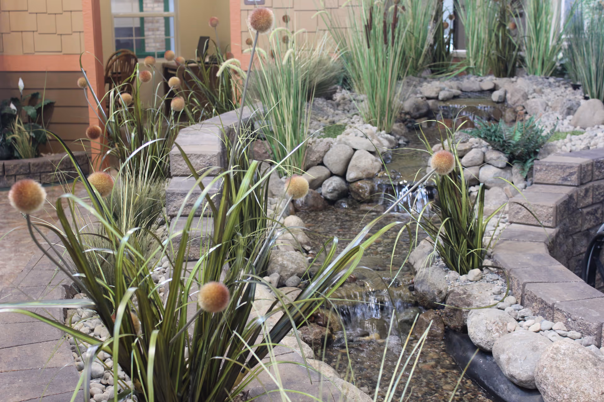Indoor water feature with a small flowing stream surrounded by rocks and tall green plants with round brown flower heads, set within a facility interior with brick and stone walls.