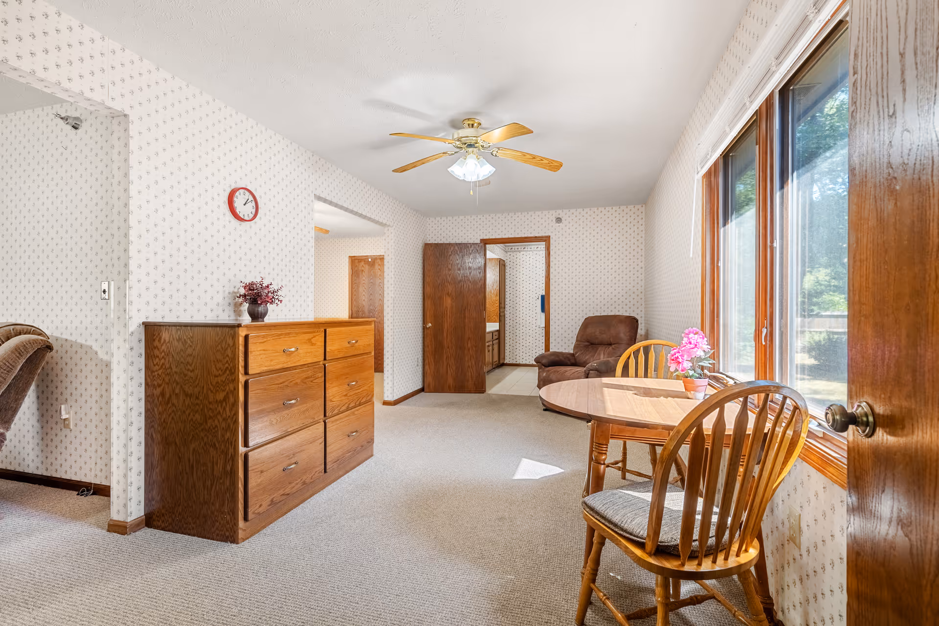 Bright living room with a wooden dining table and chairs by a large window, a wooden dresser, recliner, and ceiling fan.