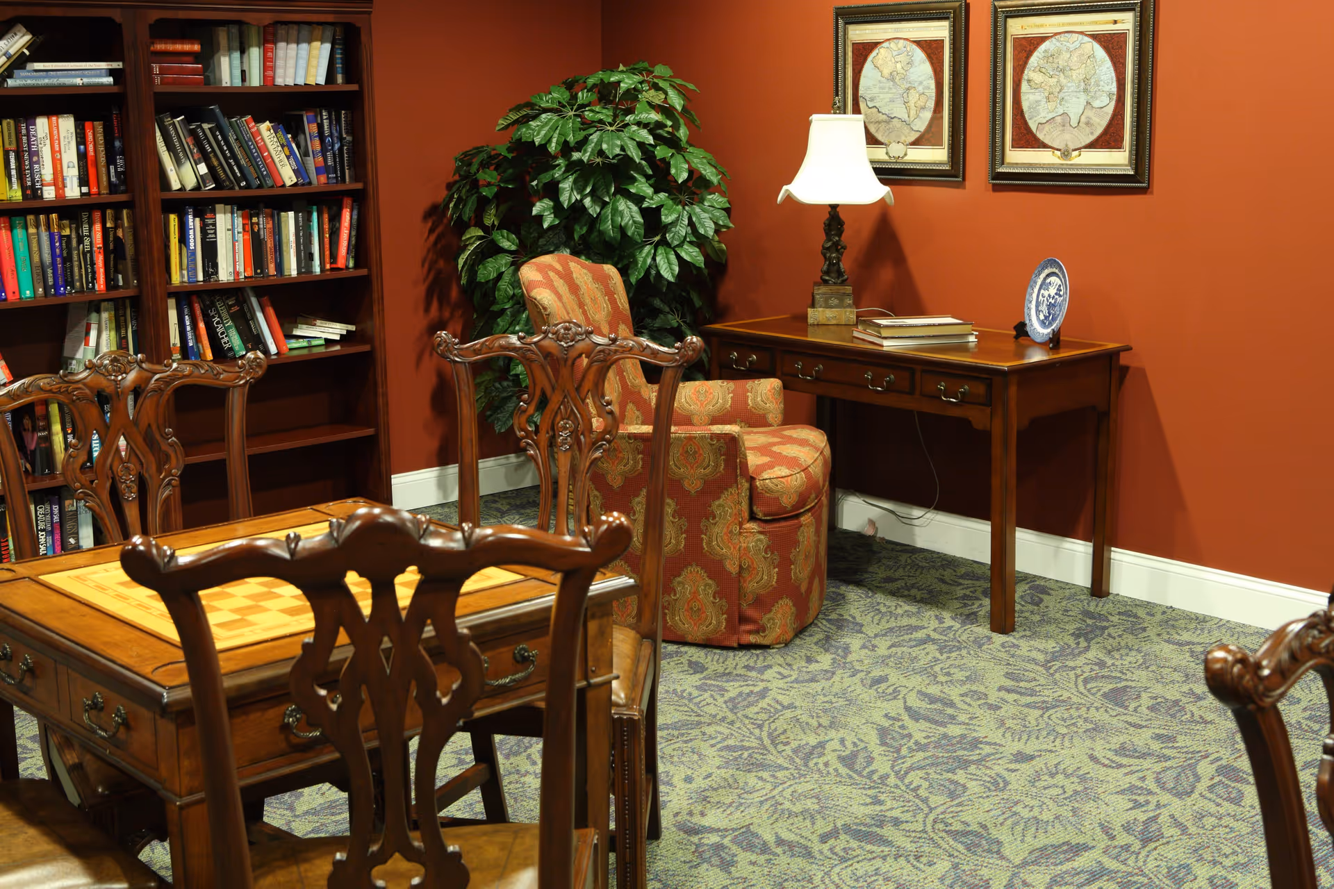 A cozy senior living room with a wooden table featuring a built-in chessboard, surrounded by ornate wooden chairs. In the background, there is a bookshelf filled with books, a patterned armchair, a wooden desk with a table lamp, some books, and a decorative plate. Two framed vintage world maps hang on the red wall, and a large green plant adds a touch of nature to the room. The floor is covered with a patterned carpet.