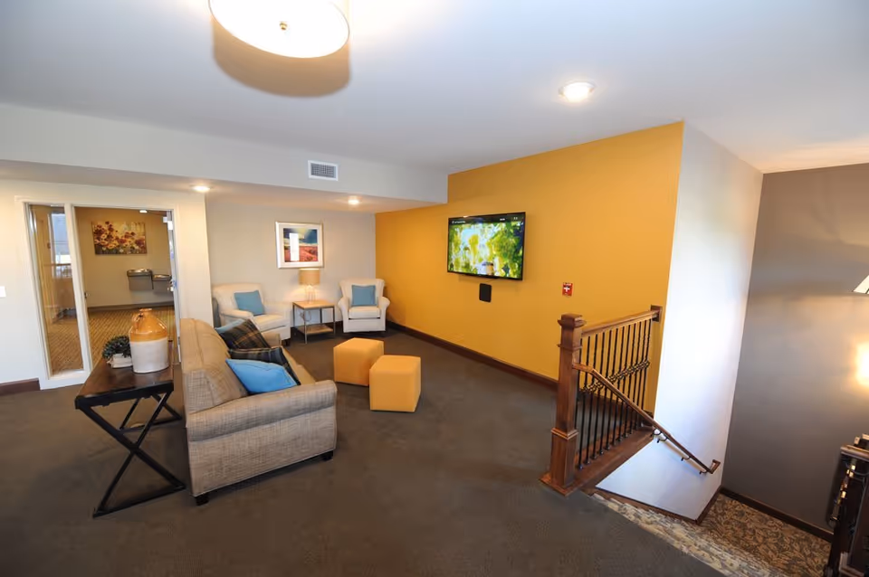 A cozy living room area with a beige sofa, two white armchairs with blue cushions, two yellow ottomans, a wall-mounted TV on a mustard yellow accent wall, and a wooden staircase with black iron railings leading downstairs. There is a side table with a lamp between the armchairs and a console table with decorative items behind the sofa. The room has carpeted flooring and neutral-colored walls.